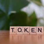 Close-up of Scrabble tiles spelling 'Token' on a wooden surface with a blurred green background.